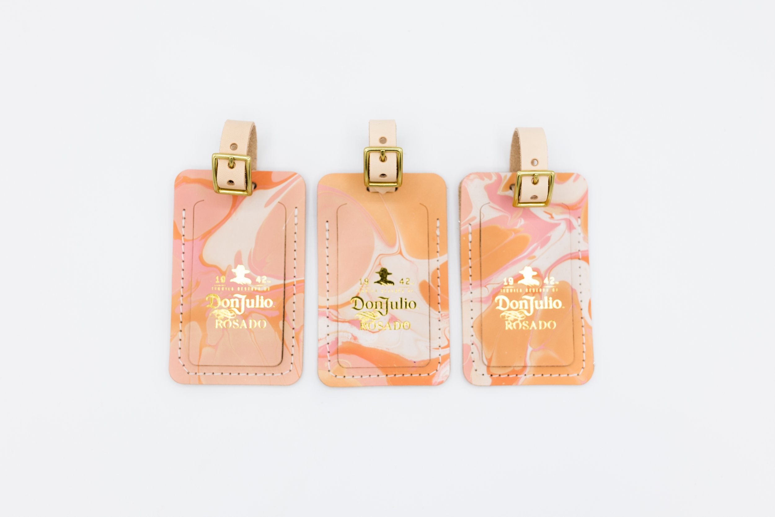 Three Don Julio Rosado branded luggage tags with pink and orange swirled patterns and gold embossing, featuring beige straps with gold buckles, on a white background.
