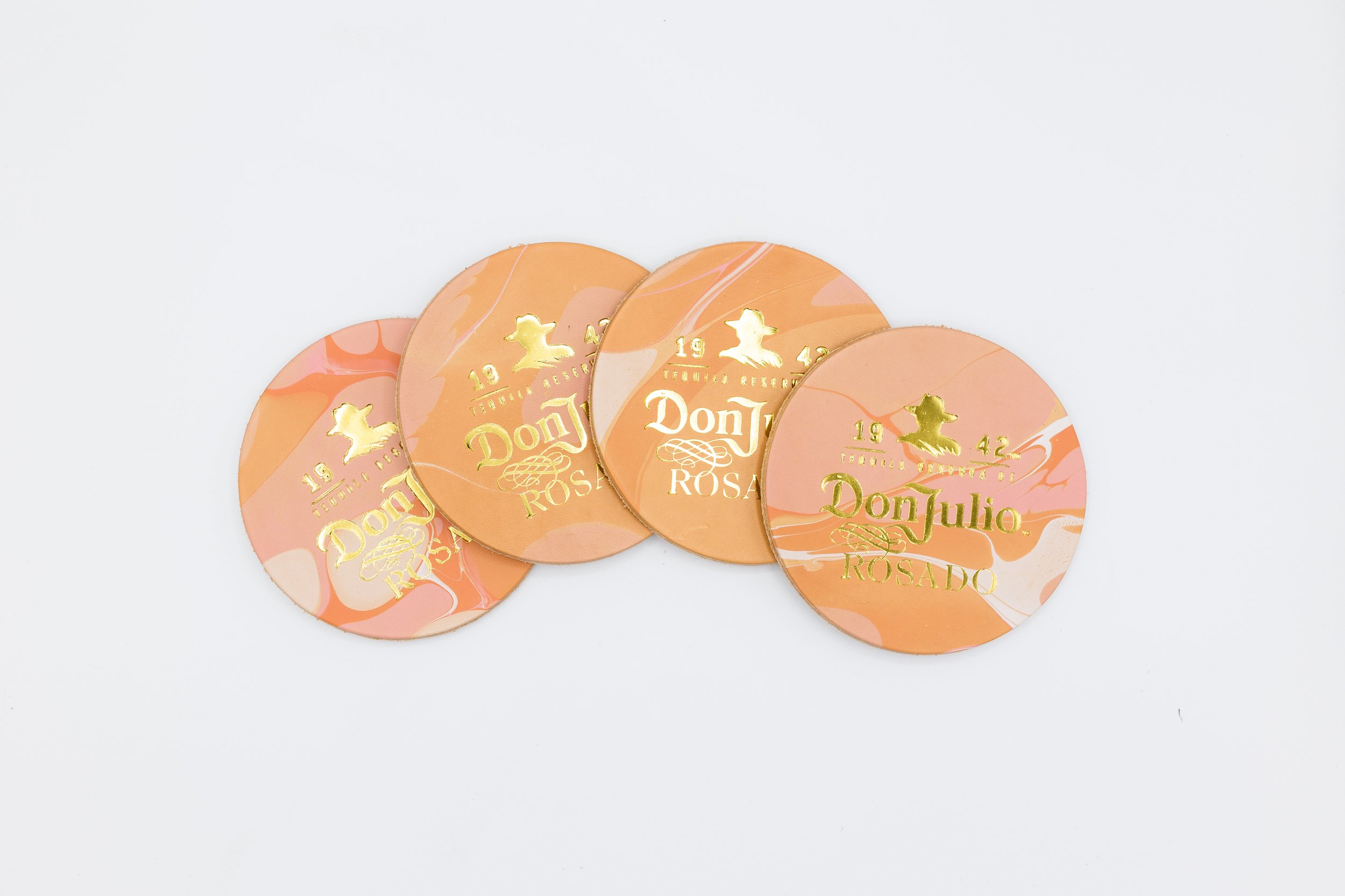 Four round peach-colored coasters with gold lettering saying "Don Julio Rosado 1942," featuring a stylized logo.