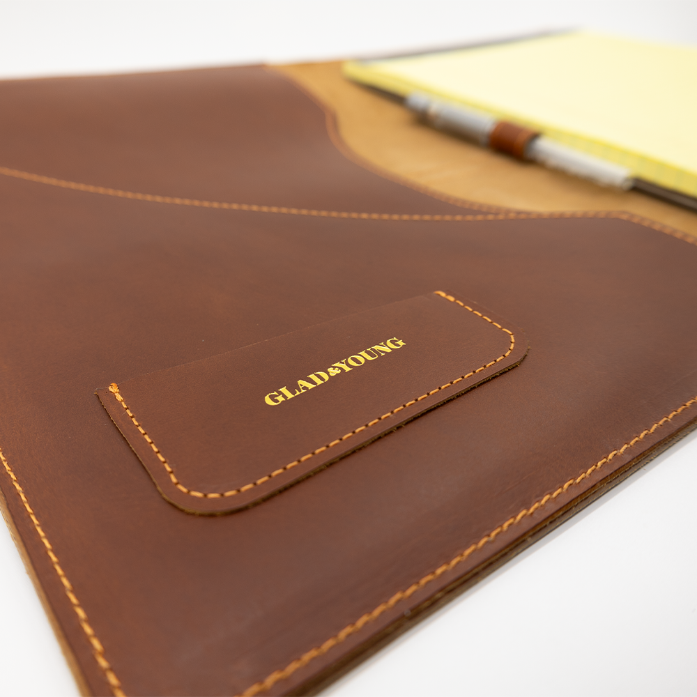 Brown leather portfolio with embossed "GLAD & YOUNG" inscription, yellow stitching, and a notepad partially visible inside.