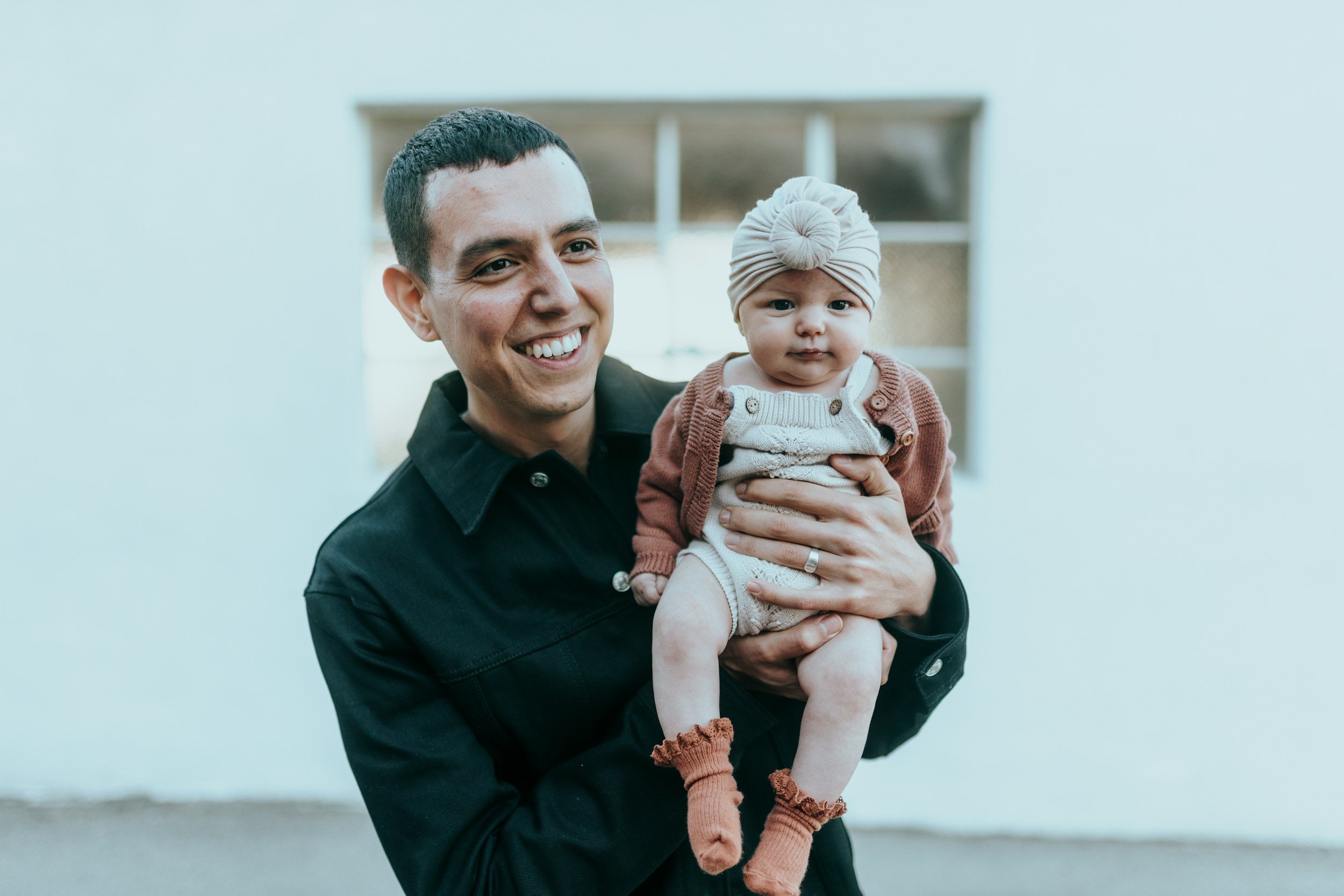 A smiling man holding a young baby girl with a beige headwrap and brown socks, standing in front of a white wall with a window.