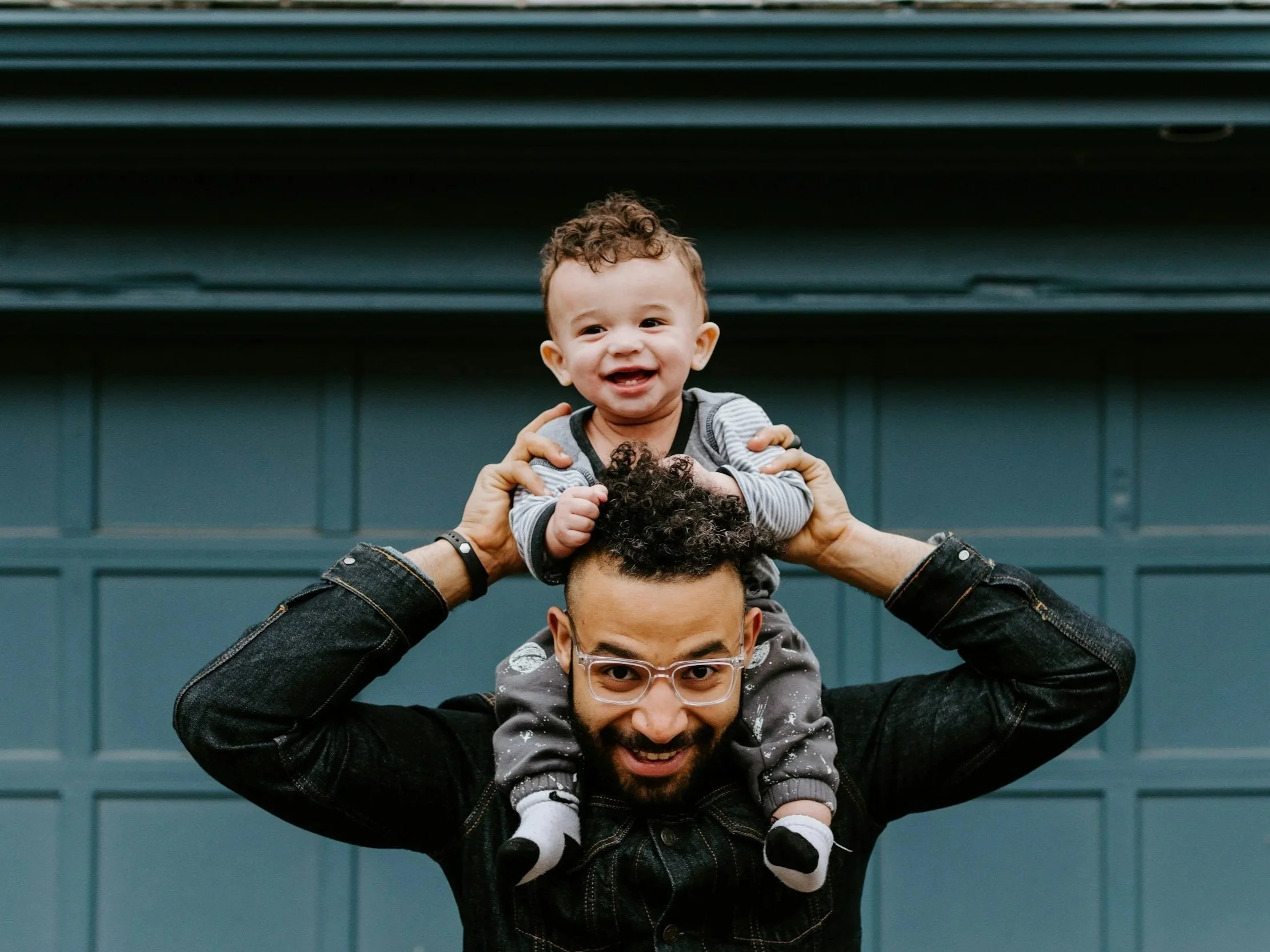 A man with glasses and a beard is smiling and carrying a young child on his shoulders. The child is smiling, has curly hair, and is wearing a striped shirt.