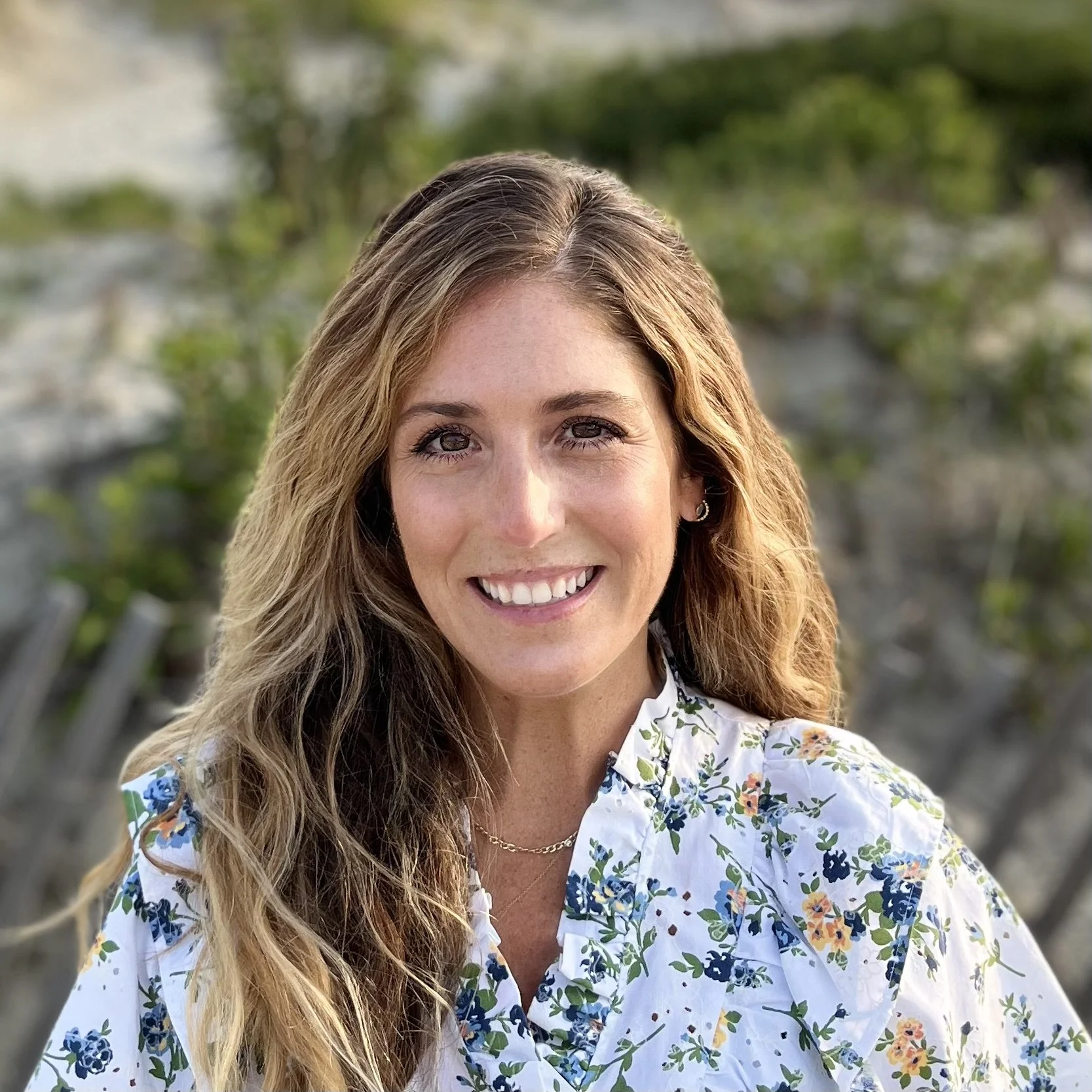 A smiling woman with long wavy blond hair, wearing a white blouse with blue and orange floral patterns, outdoors with greenery in the background.