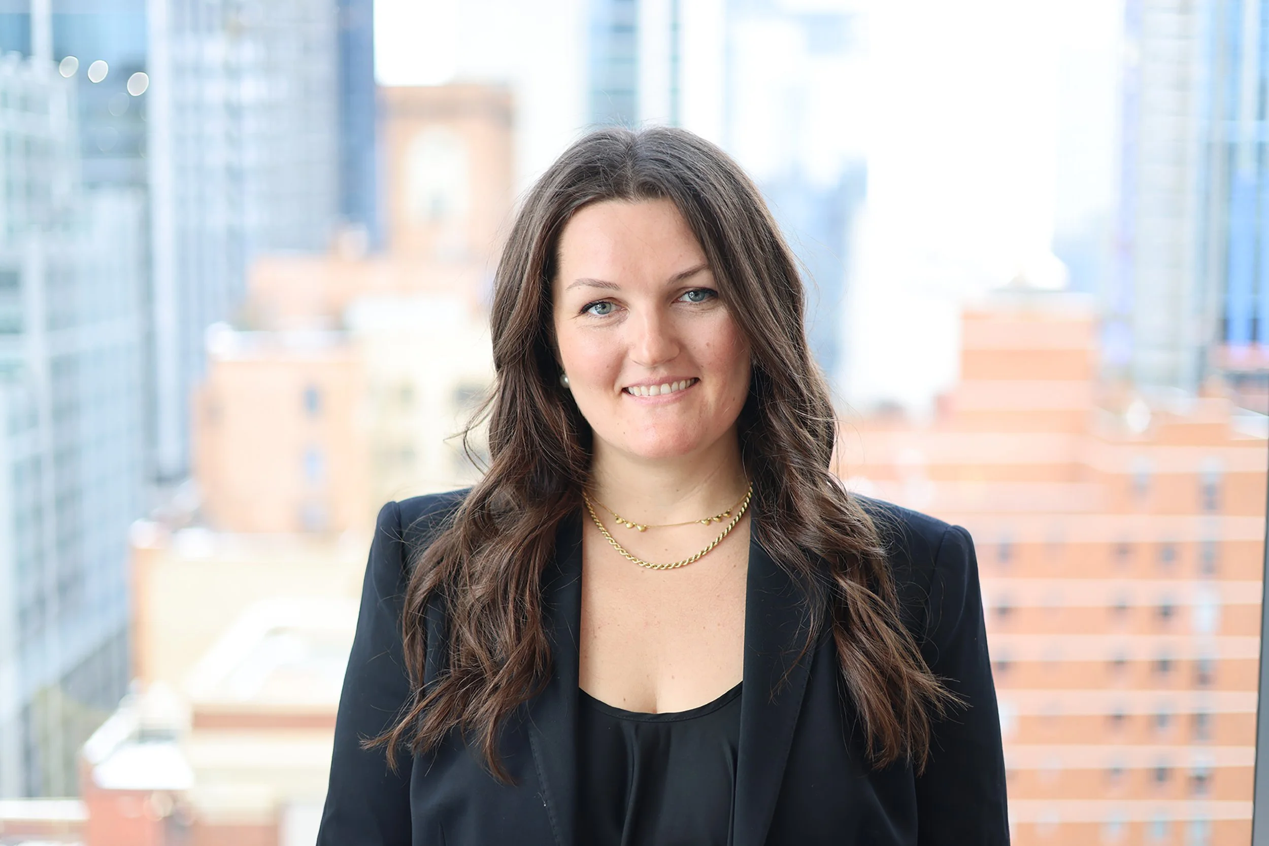 A woman with long brown hair wearing a black blazer and gold necklaces, smiling in front of a cityscape background with tall glass buildings.