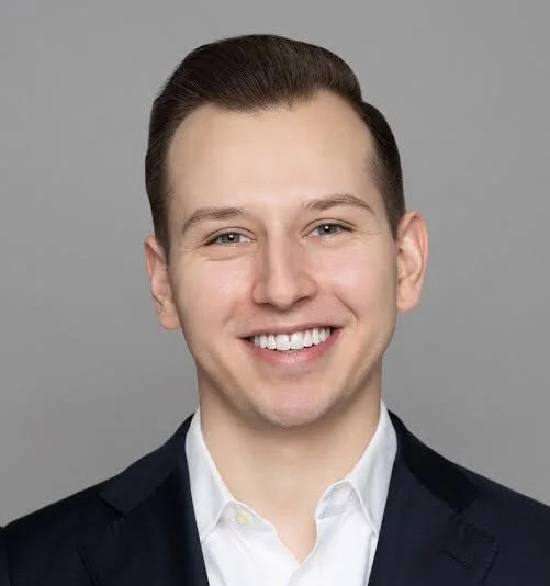 A young man with short, dark hair smiling, dressed in a dark blazer and white shirt against a light gray background.
