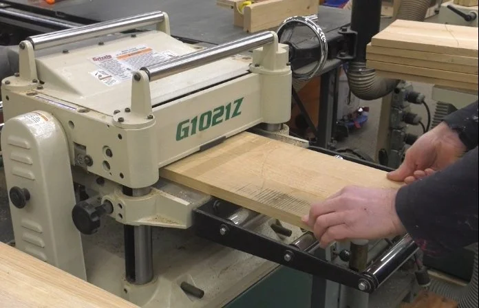 running a board of white oak through a planer, with a stack of more boards in the background