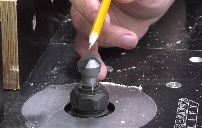 a hand using a pencil to point at the tip of a v-groove router bit that is installed in a router table