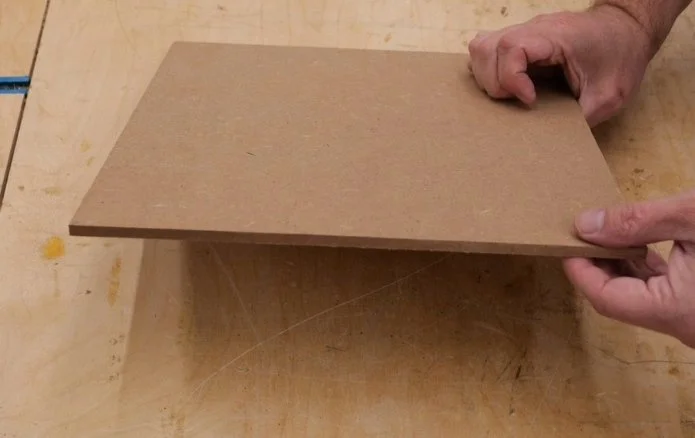 a 12 inches by 12 inches square piece of MDF that is 1/4" thick being held at an angle by hands on a wooden workbench top