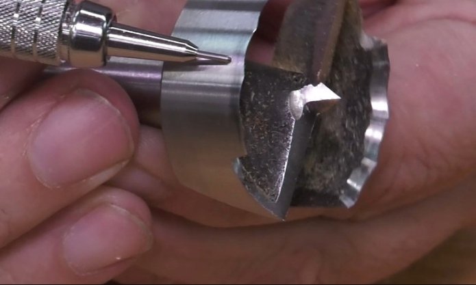 fingers holding a forstner bit on it's side while a mechanical pencil points to the edge of the bit