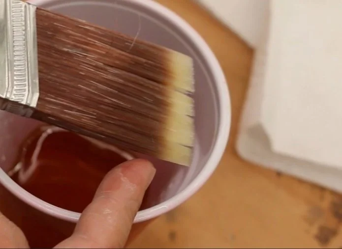 a finger pointing at the bottom half of a paint brush that is wet with an oil based finish, with a red solo cup that has oil based finish in it, blurred in the background