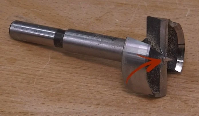 a digital red arrow pointing to the center spur of a forstner bit lying on a wooden benchtop