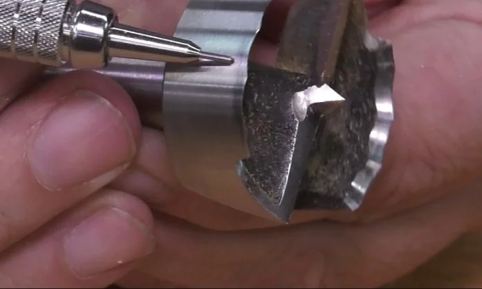 fingers holding a forstner bit on it's side while a mechanical pencil points to the edge of the bit