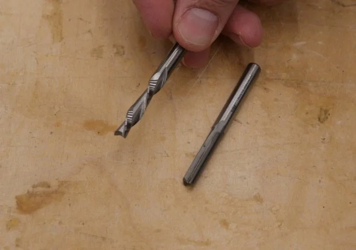 1/4 spiral bit being held up by fingers, and a 1/4 inch straight bit laying on a wooden table top