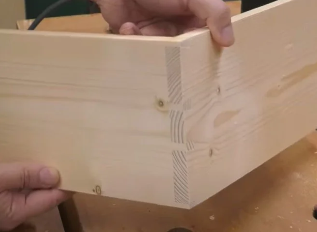 Person assembling a piece of light-colored wooden furniture, possibly a drawer or box, in a workshop setting.