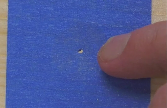 a piece of blue tape covering a hole in wood with a  small hole pocked through the center of the tape