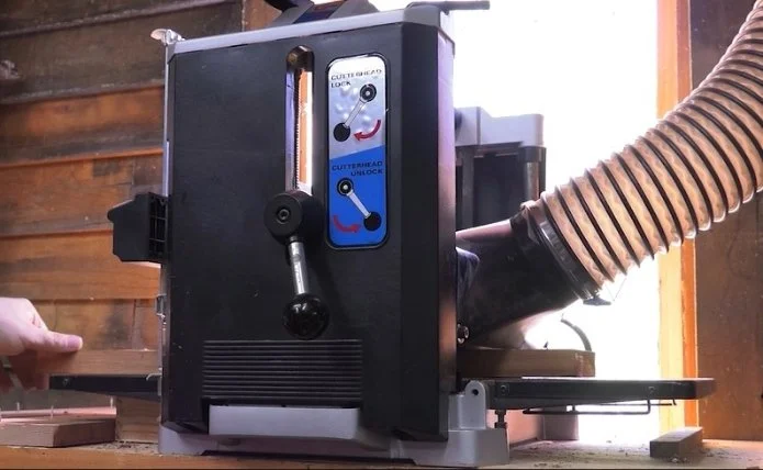 a hand guiding the end of a piece of wood into a thickness planer sitting on a table top with a window and wooden wall in the background