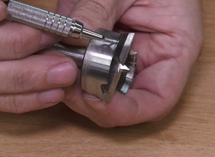 the tip of a mechanical pencil pointing at the continuous rim of a forstner bit being held sidesway by a pair of hands