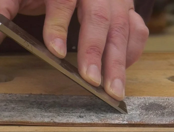 a hand on the back side of a chisel, running the edge of the bevel along a leather strop covered in abrasive paste