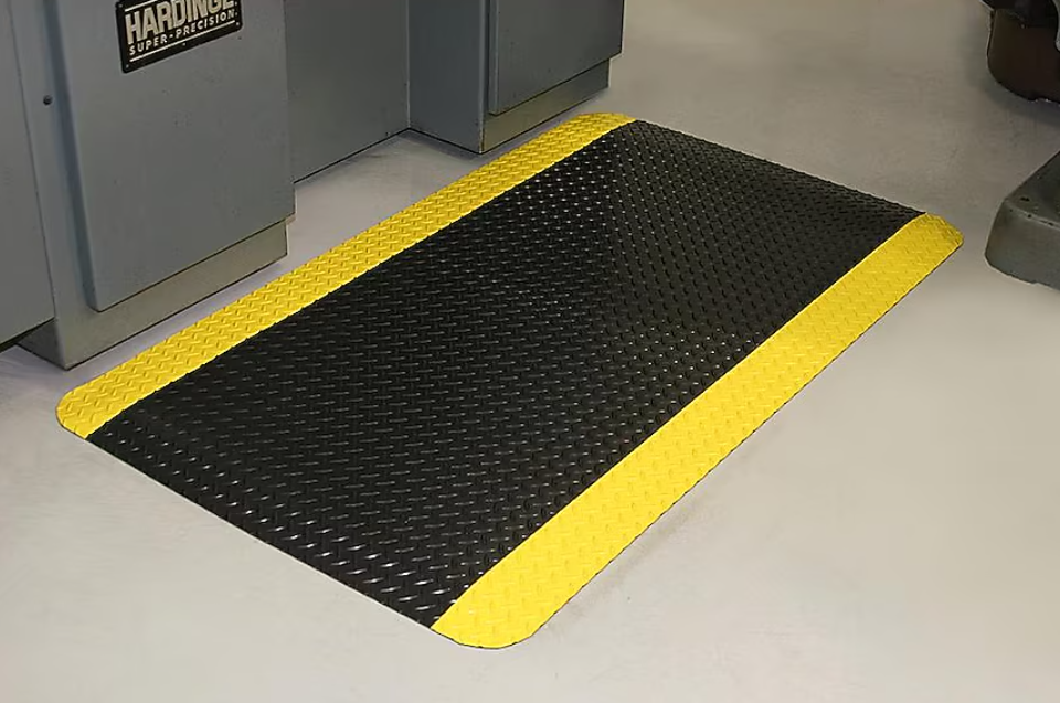 Floor mat with black rubber surface and yellow border in a carpeted workshop