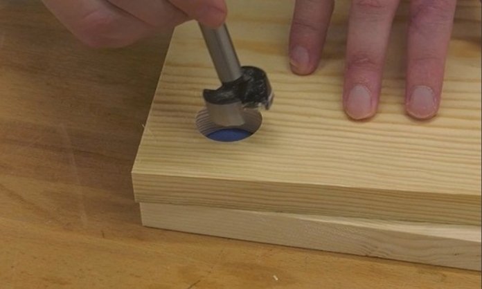 hovering a forstner bit over two pieces of wood, the top piece having a hole cut out that matches the size of the bit, and the piece below with blue tape on it that can be seen through the hole in the top piece of wood. on top of a wooden bench top