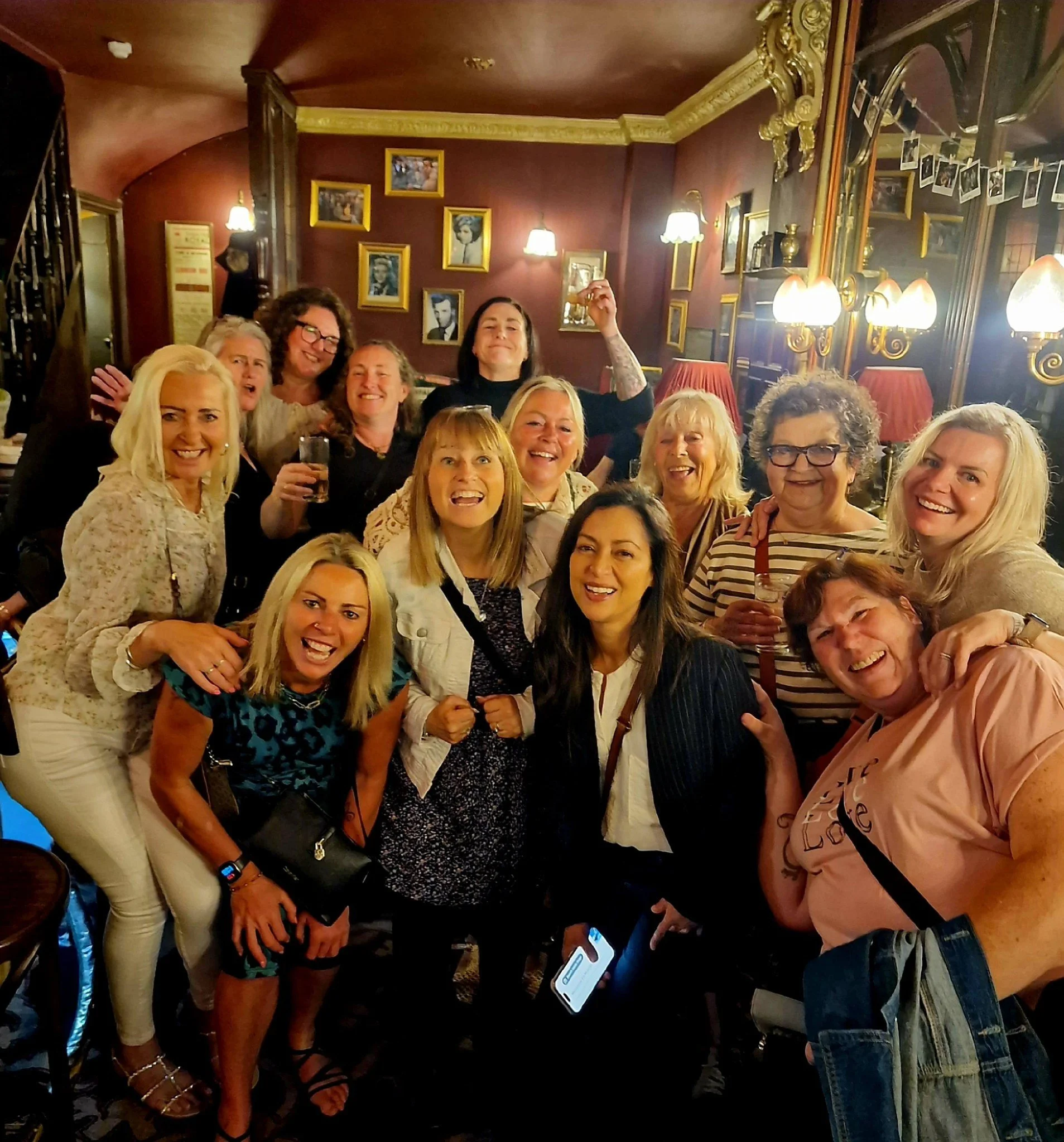 Group of women smiling and celebrating in a cozy, dimly lit restaurant or bar with framed pictures on the wall and vintage lamps.