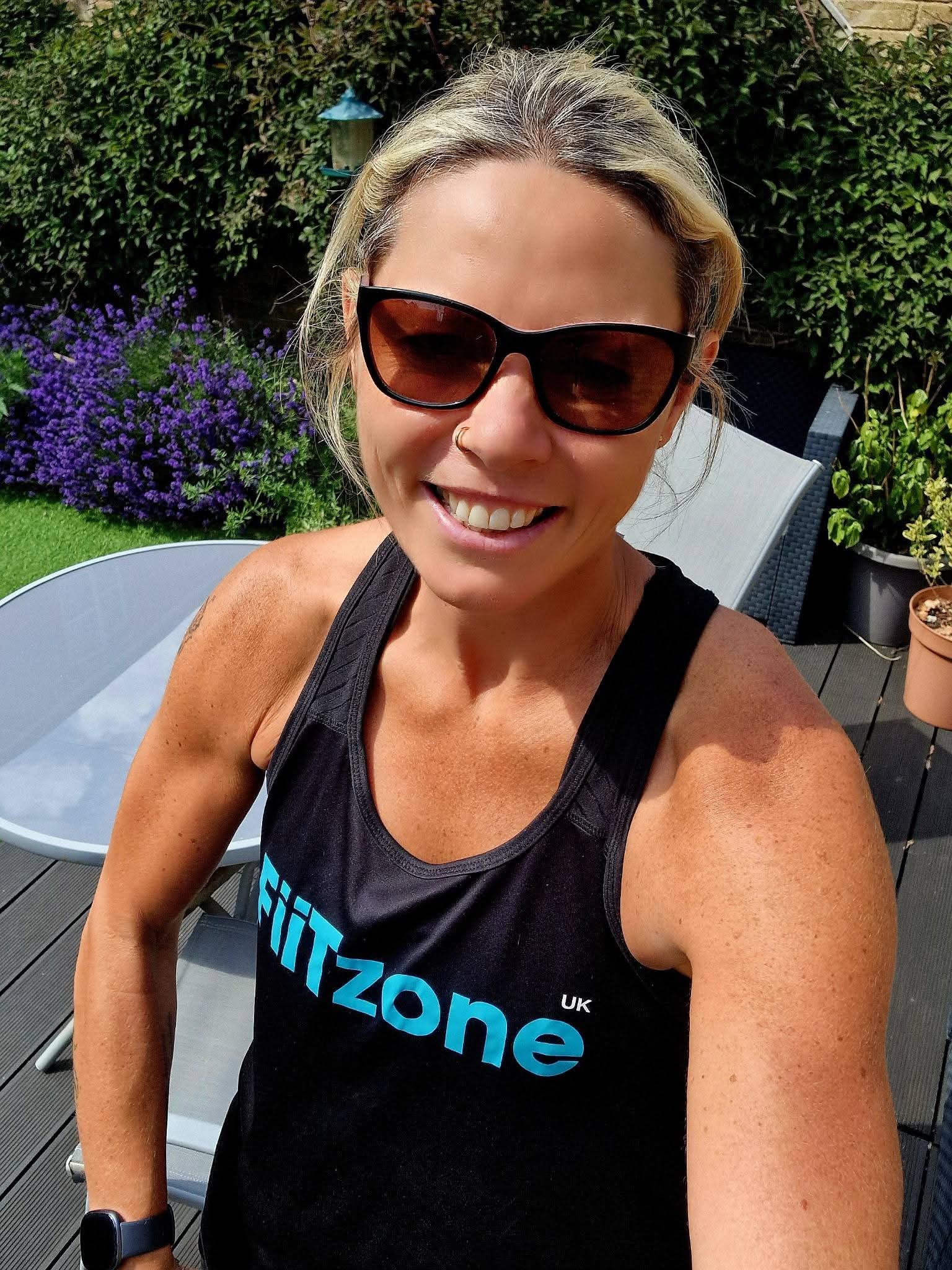 A woman wearing sunglasses, a black fitness tank top with 'FITZONE UK' written on it, taking a selfie outdoors in a garden with purple flowers, green bushes, and patio furniture.