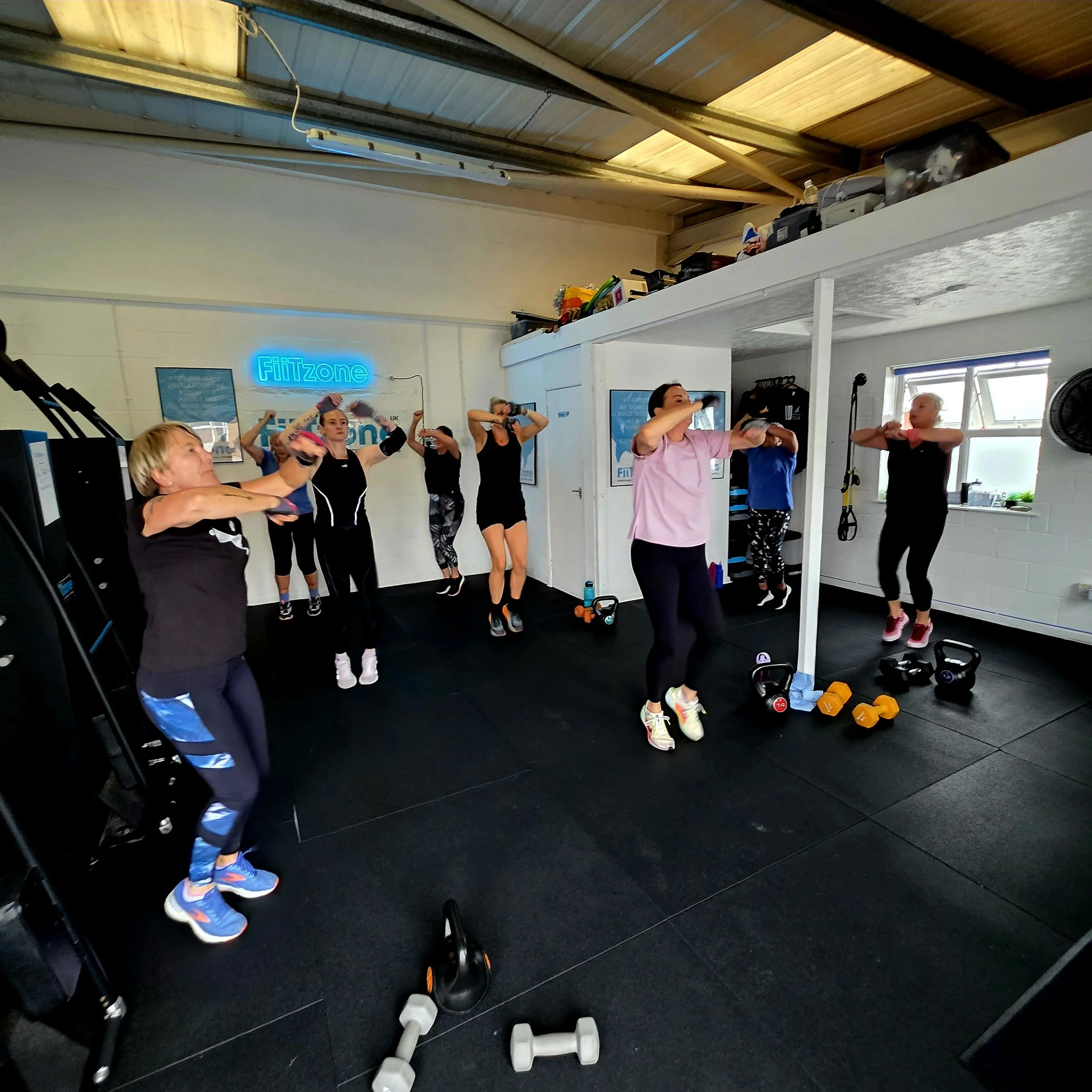 Group of women participating in a fitness class inside a gym, performing arm exercises, with various workout equipment and a neon sign reading 'FitZone' in the background.