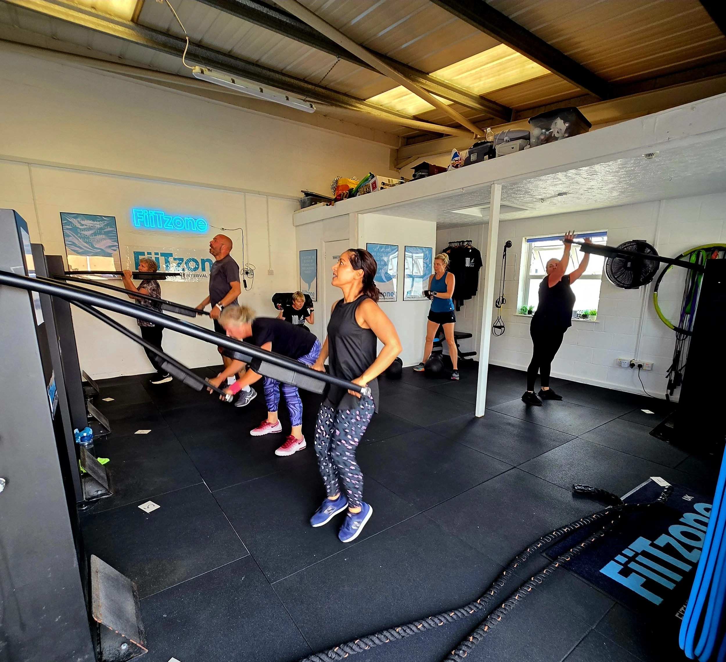 People exercising on treadmills and engaging in activities in a gym with a wooden ceiling and a sign that reads 'Fitzone'.