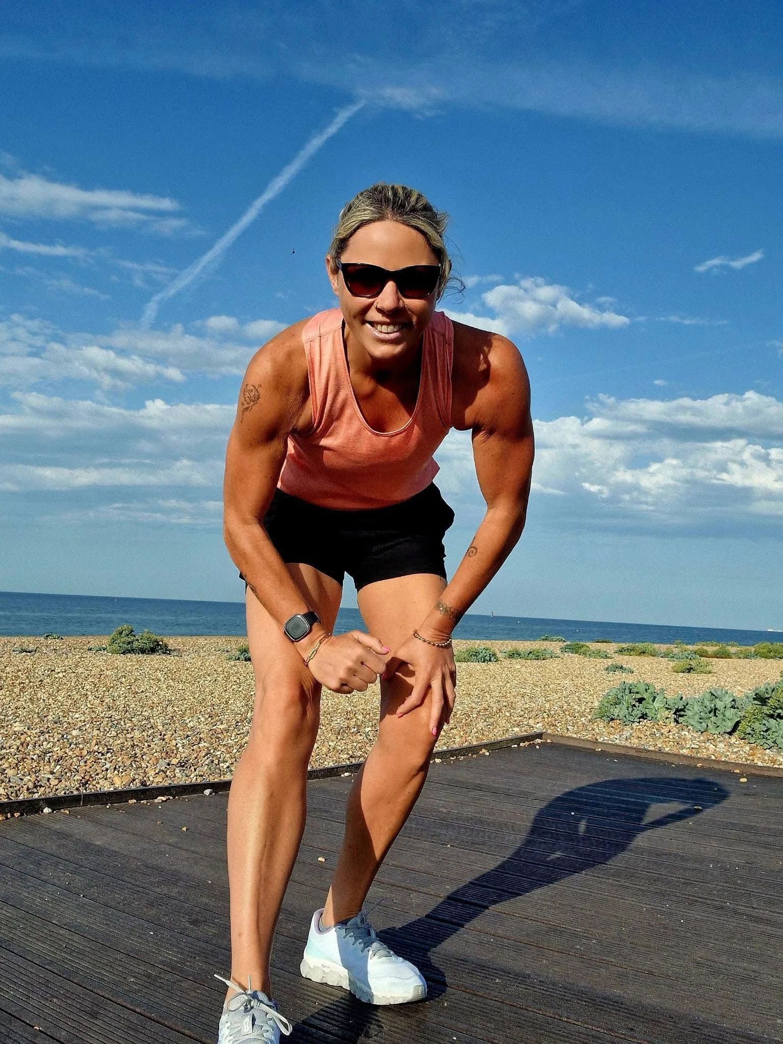 Woman in athletic wear crouching outdoors on a wooden path near the beach, wearing sunglasses, a pink tank top, black shorts, and white sneakers, with the sky and ocean in the background.