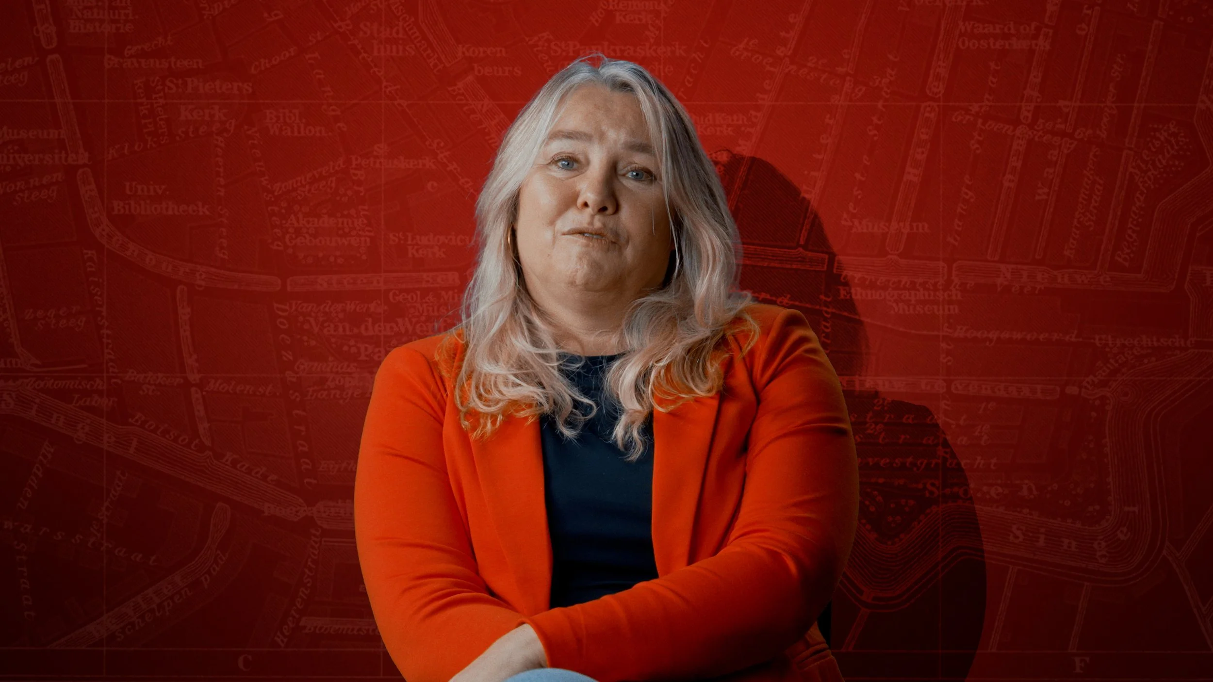 Vrouw met lange blonde haar in een oranje jas en een zwarte top zittend voor een rode achtergrond met een kaart of plattegrond.