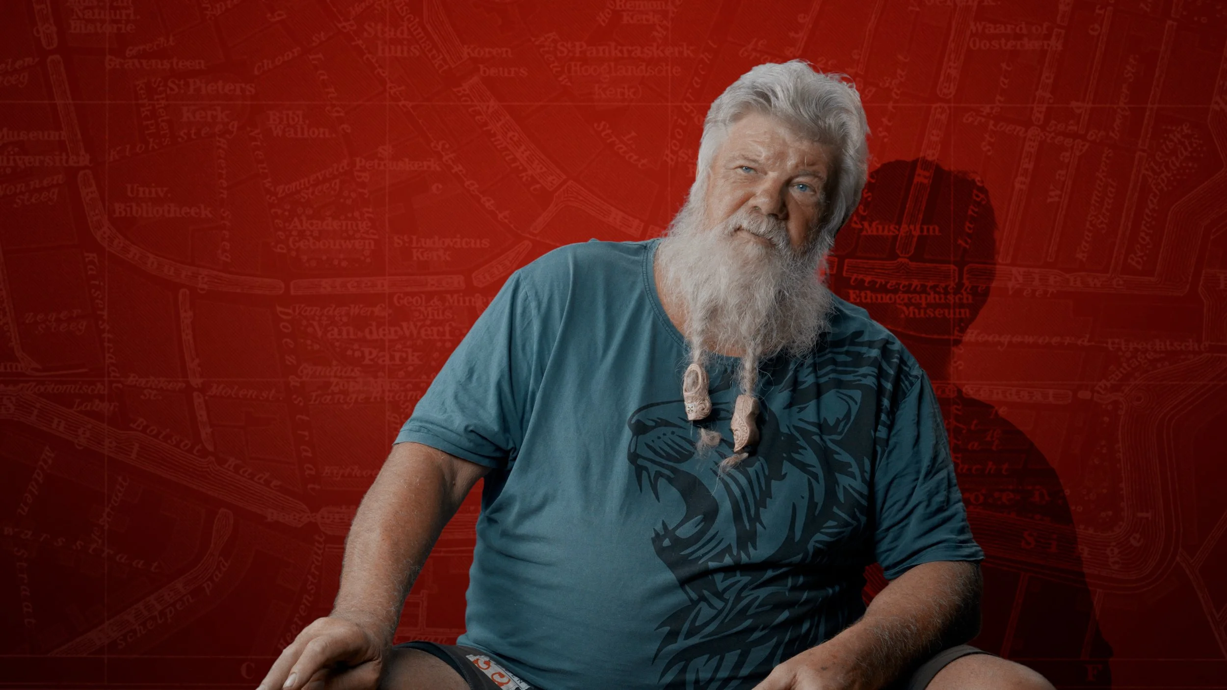 Een oudere man met een lange witte baard en grijze haar, zittend tegen een rood achtergrond met een stadsplattegrond en een hartvormige locatie met de tekst "Museum Ethnographisch Museum". Hij draagt een blauwe t-shirt met een tatuur van een luipaard