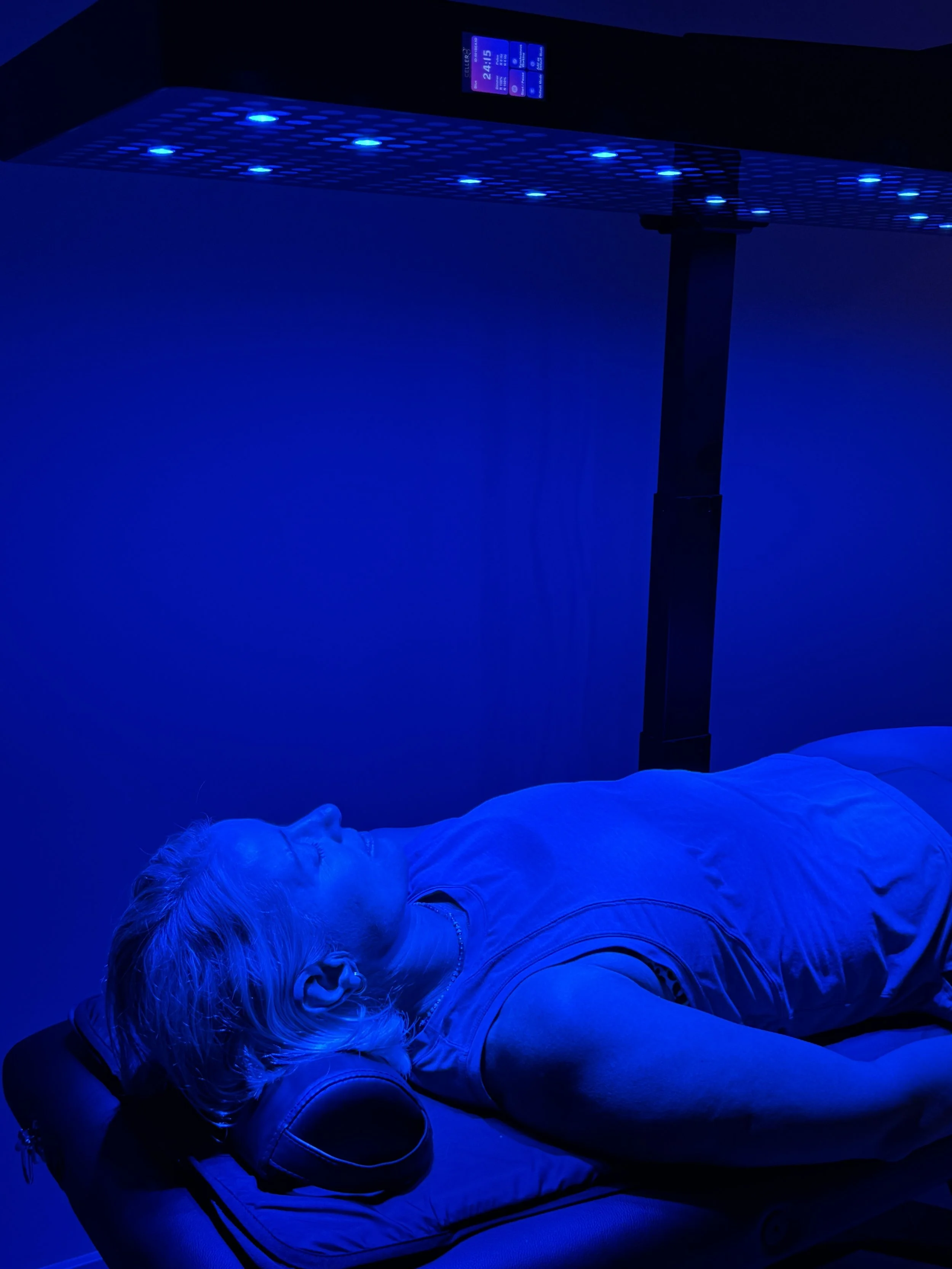 A person with blonde hair lying on a bed or reclined surface with their eyes closed, under blue LED light. There is a control panel or display on the ceiling above them.