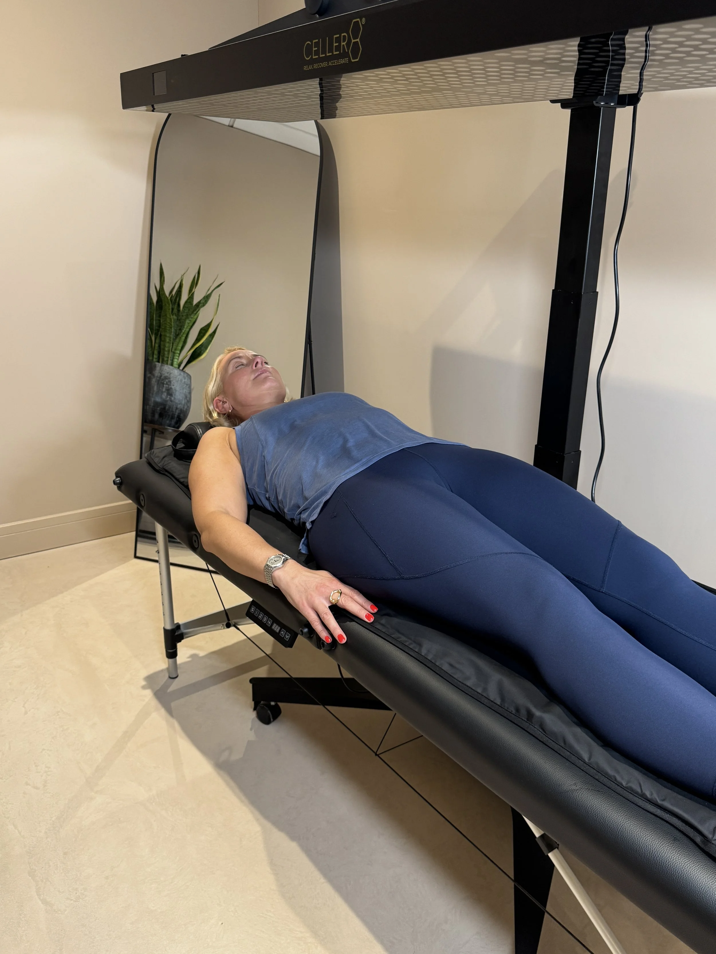 A woman lying on a therapy table under a large black and gray device labeled 'Celler,' in a room with a potted plant and a mirror.