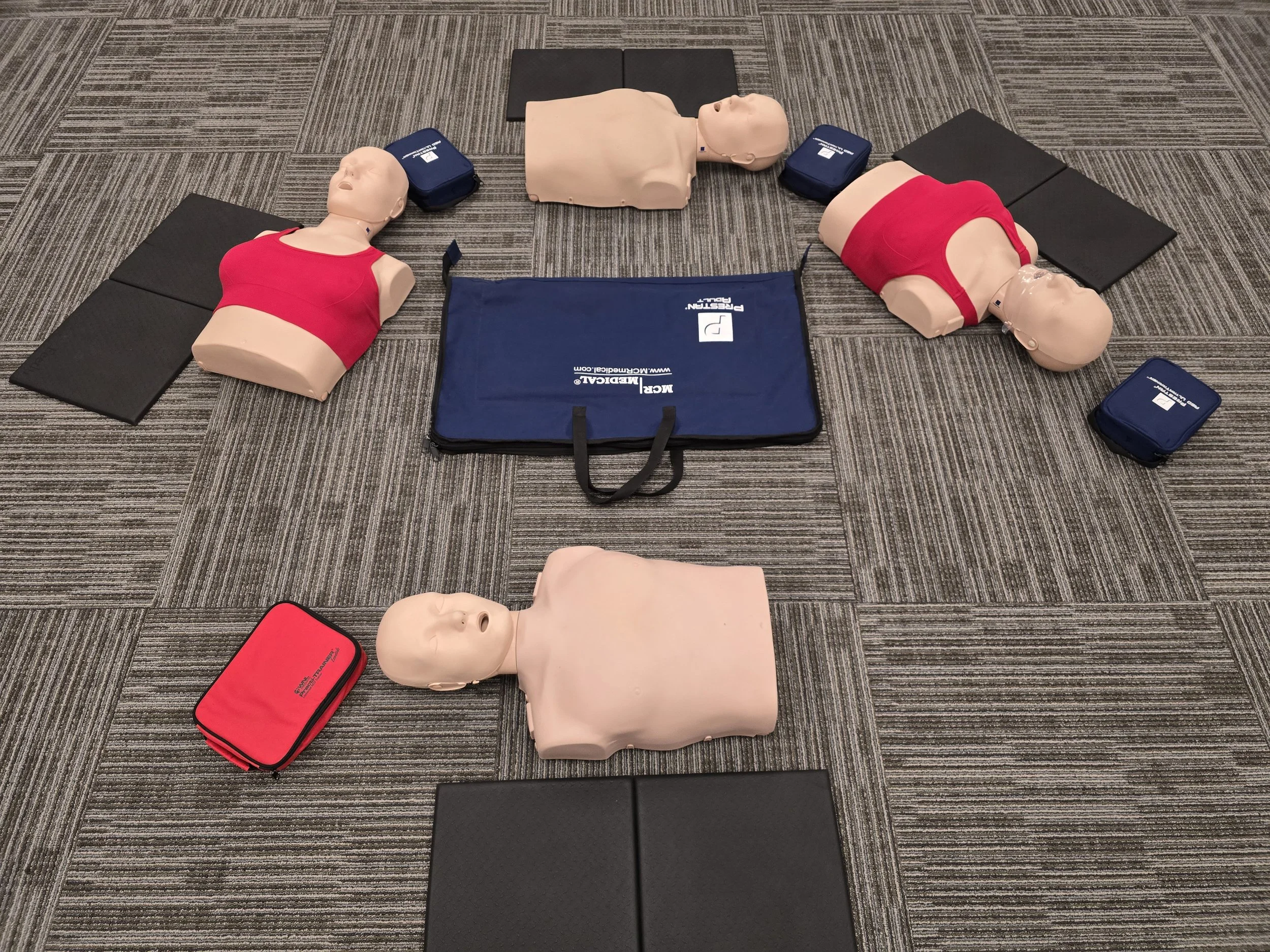 CPR training mannequins arranged in a circle on the floor with knee pads and other training items