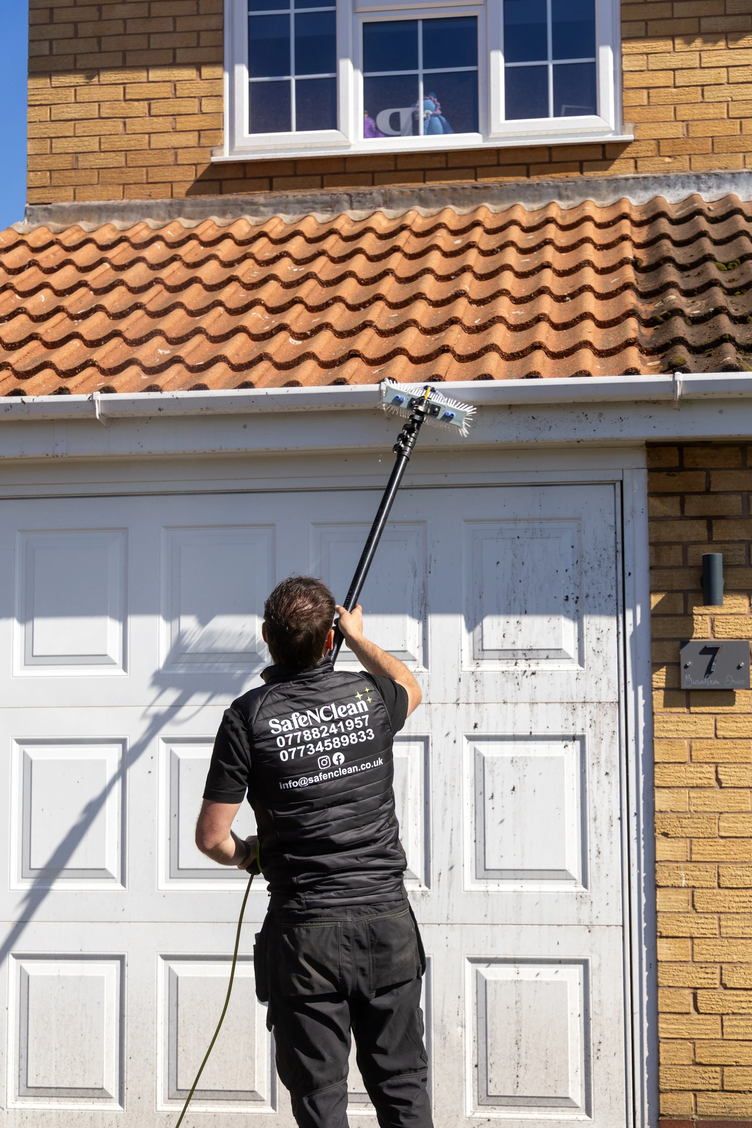 A person cleaning a white garage door with a long-handled brush, standing in front of a brick house with a tiled roof and a window above.