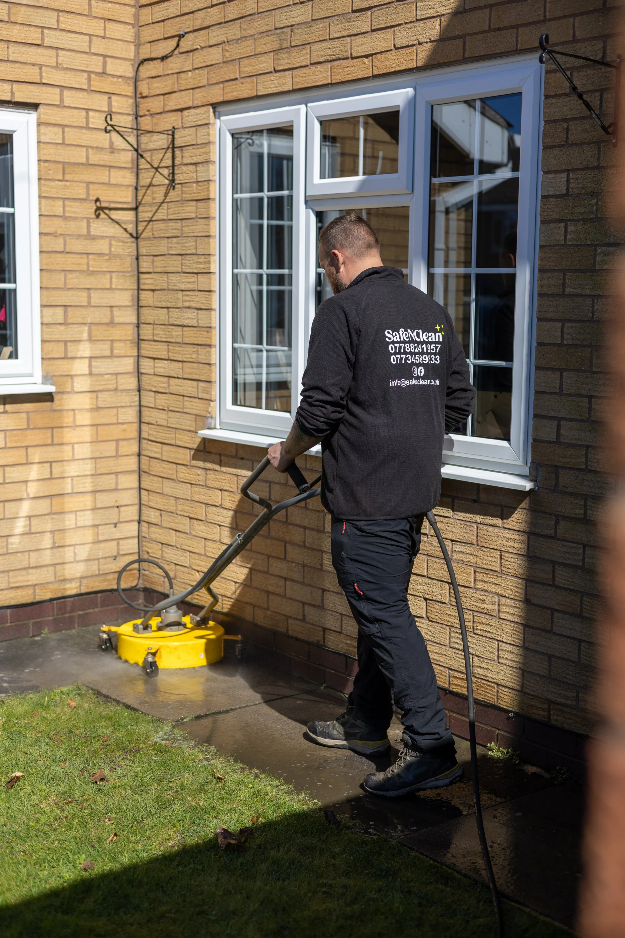 A worker pressure washing the exterior of a house with brick siding and white-framed windows.