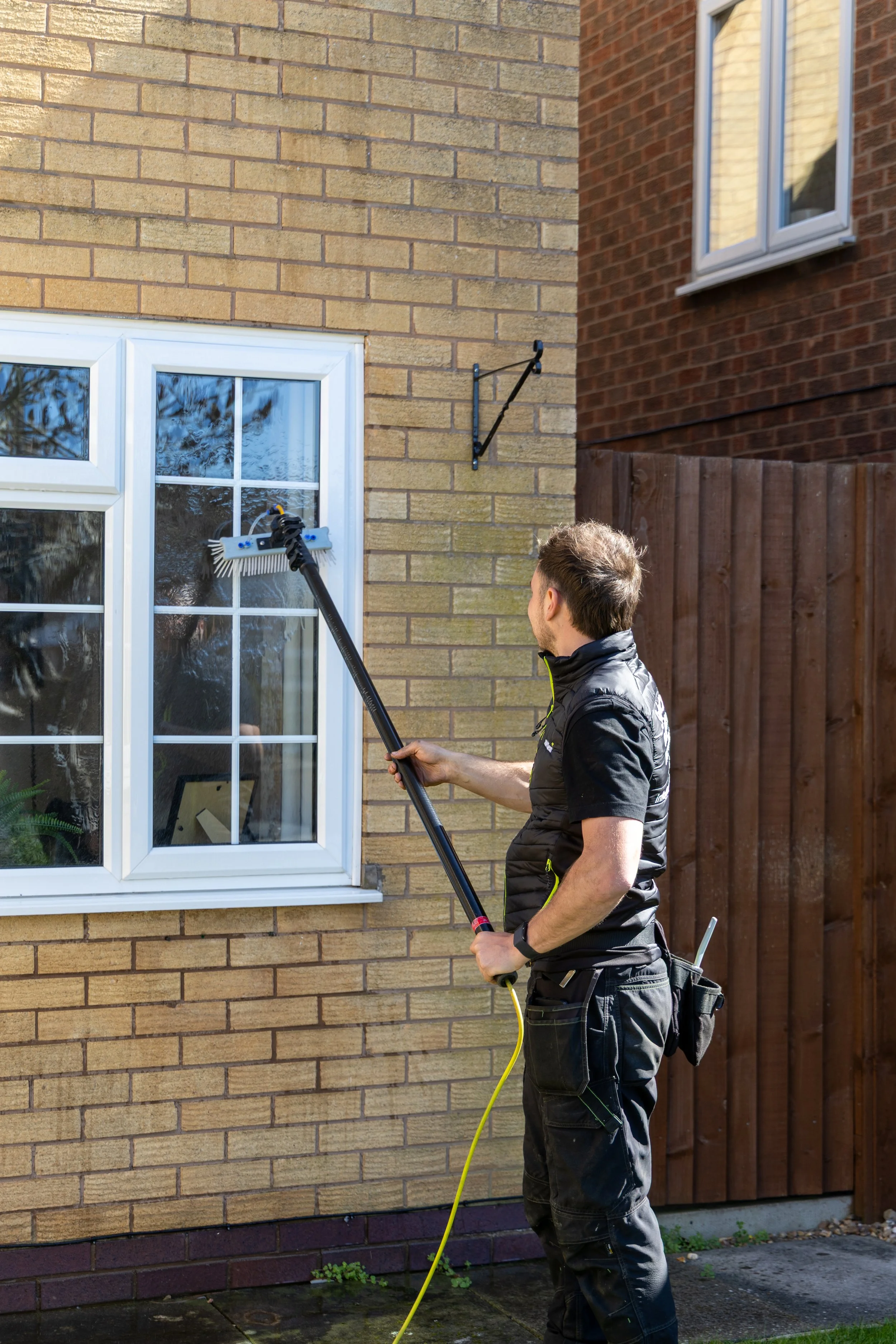 A man cleaning a window on a brick house with a pressure washer or wide brush, standing outside on a paved surface.