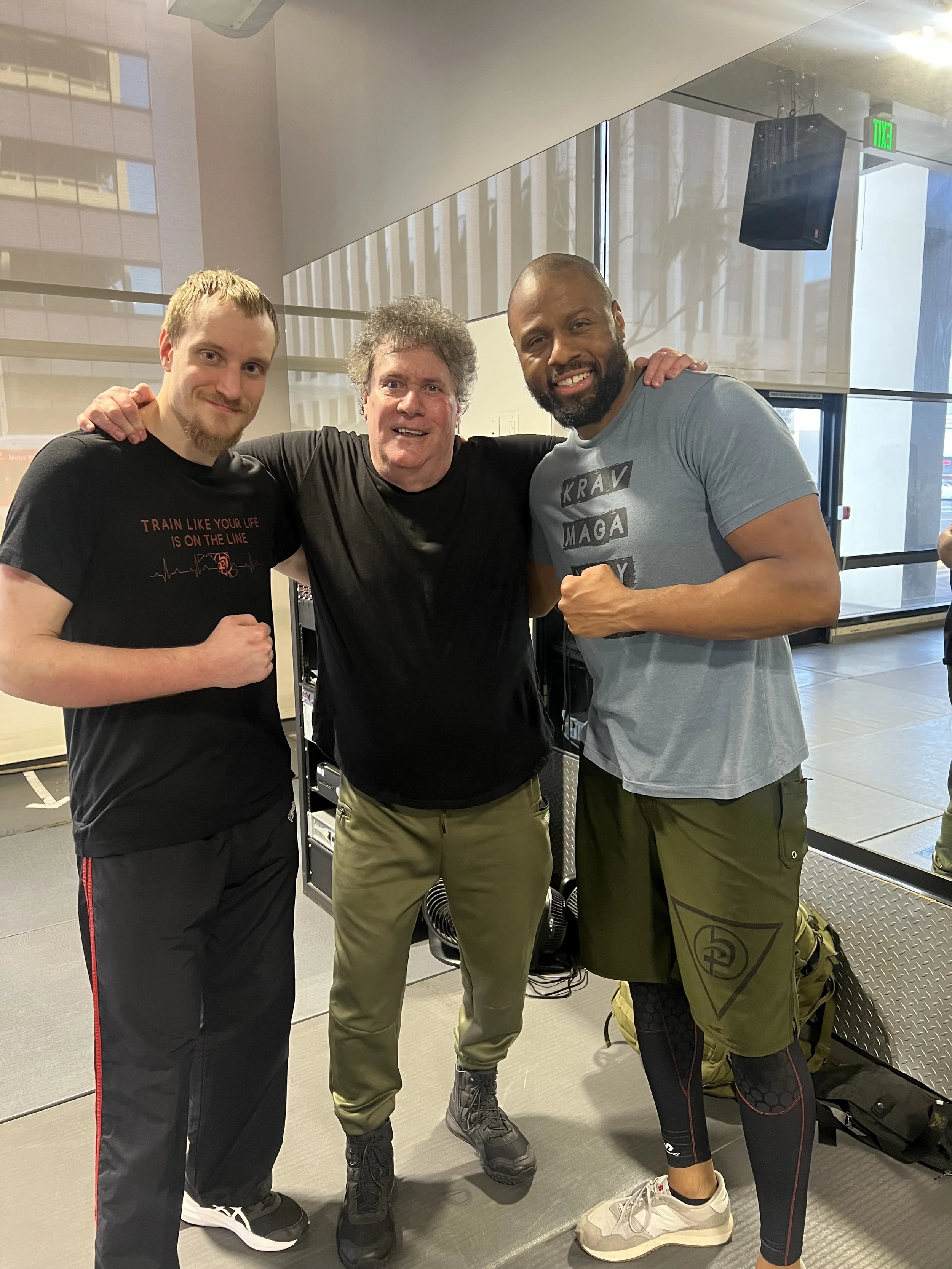 Three men standing together inside a gym, smiling at the camera with their arms around each other. The man on the left is wearing a black T-shirt with red text, the middle man is wearing a black shirt, and the man on the right is wearing a light blue T-shirt with text and shorts. They are posing confidently in a fitness setting.
