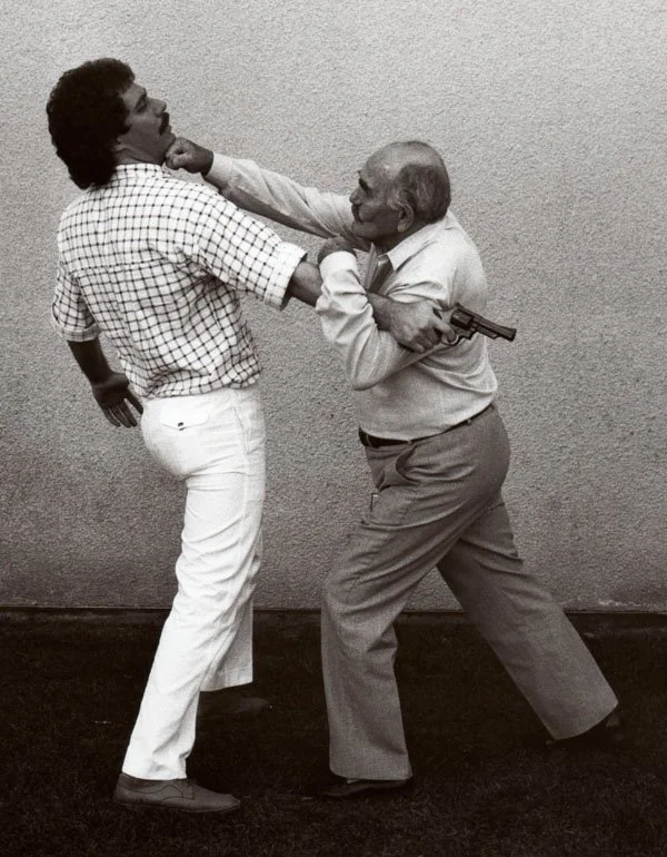 Imi Lichtenfeld and Darren Levine are engaged in a playful fight; the older man is holding a training gun and has his arm around the younger man's neck, while the younger man is holding the older man by the collar and appears to be laughing.
