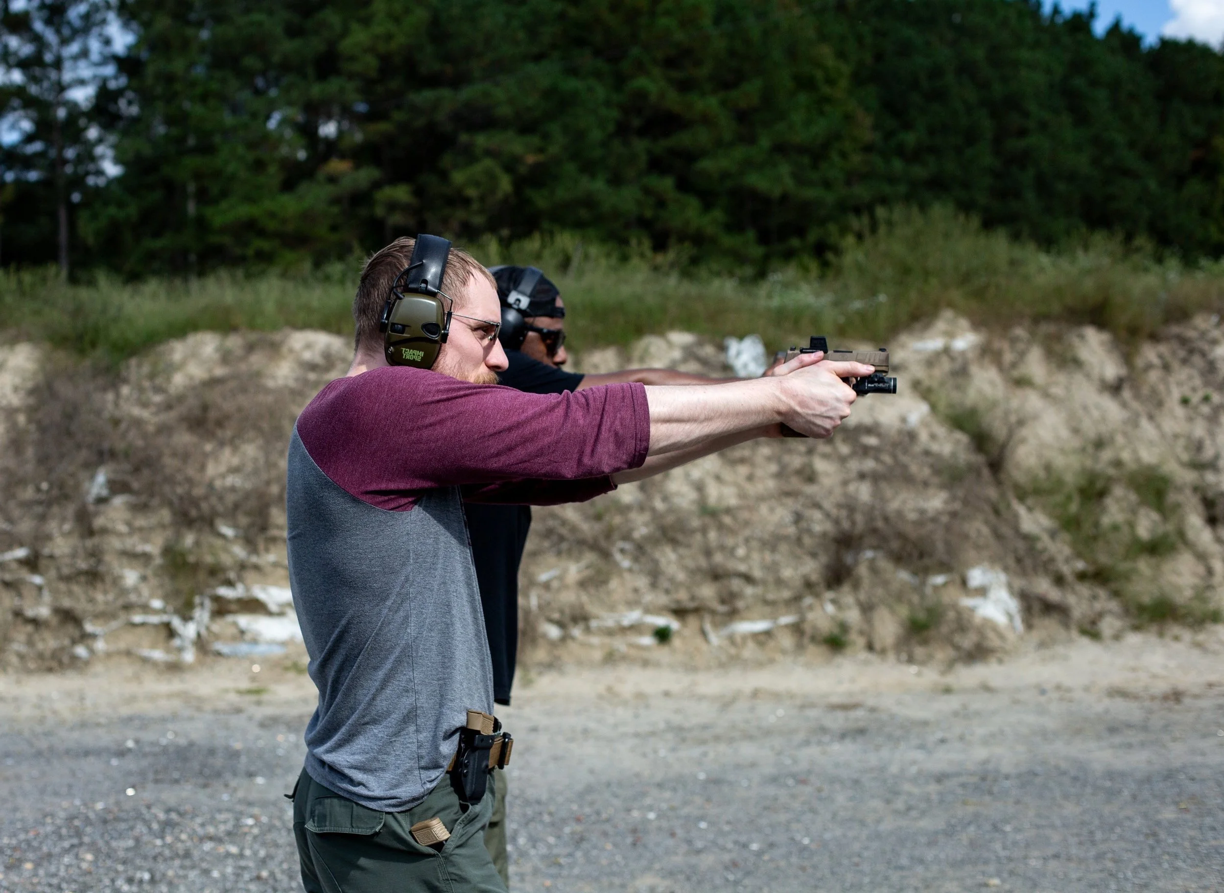 Two men wearing ear protection shooting pistols at an outdoor range.