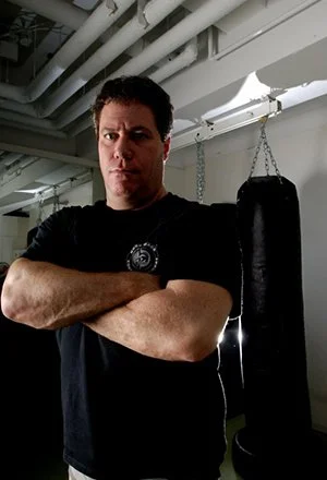 A man (Darren Levine)  with dark hair and a serious expression stands with his arms crossed in front of a punching bag in a gym. The ceiling has exposed pipes.