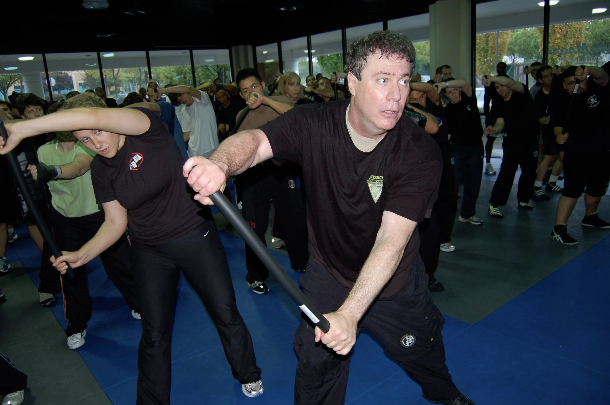 A martial arts instructor (Darren Levine) demonstrates a technique with a weapon to a group of students in a dojo with large windows and natural light.