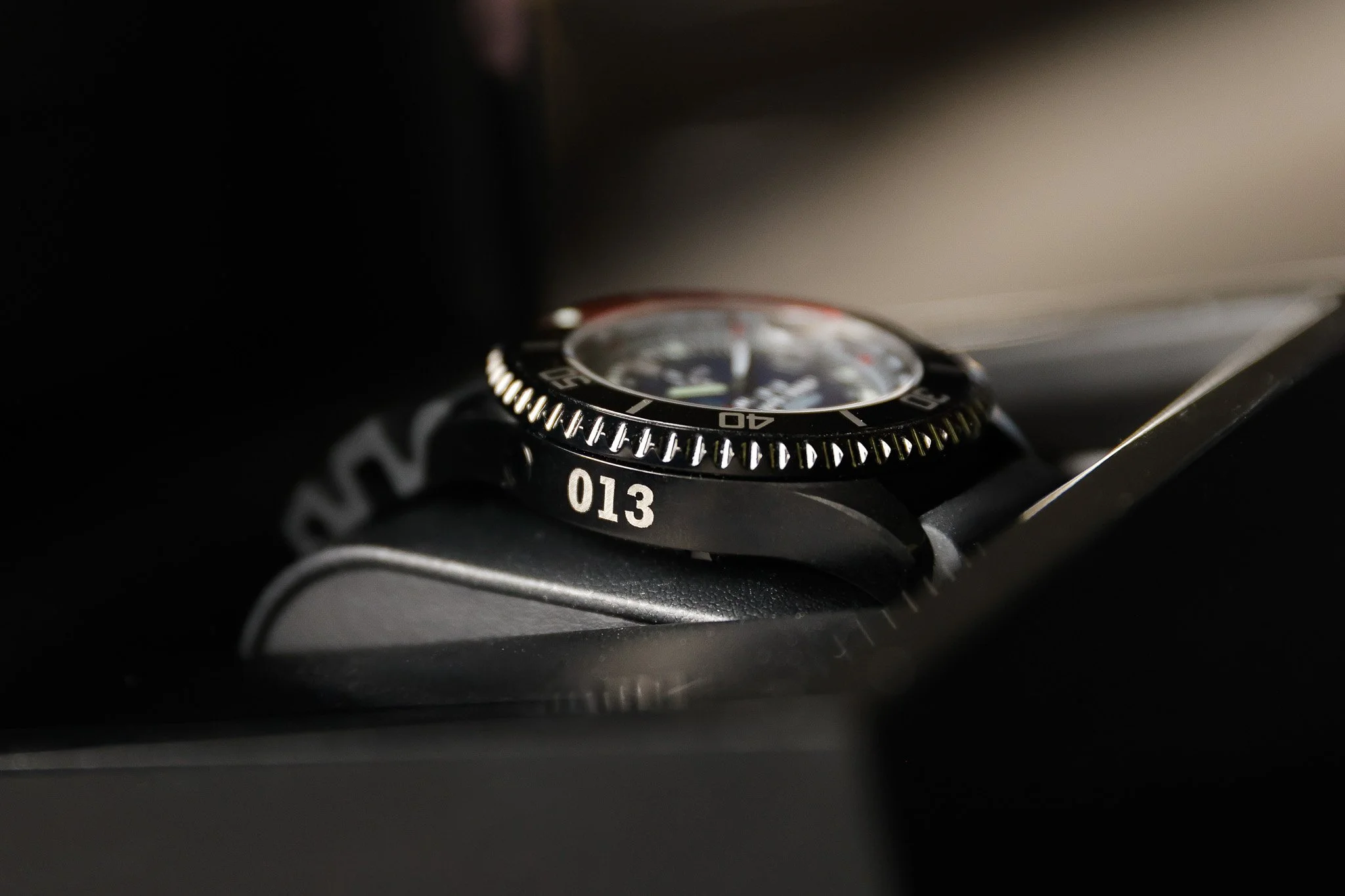 Close-up of a black analog wristwatch with a rotating bezel, placed on a surface. The watch has white markings and numbers, with the number 013 visible, and the face is slightly out of focus.
