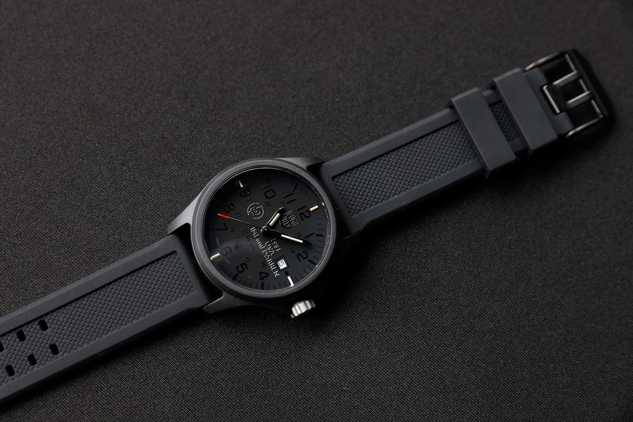 Black wristwatch with a round face, multiple dials, a date window, and a textured strap, placed on a dark surface.
