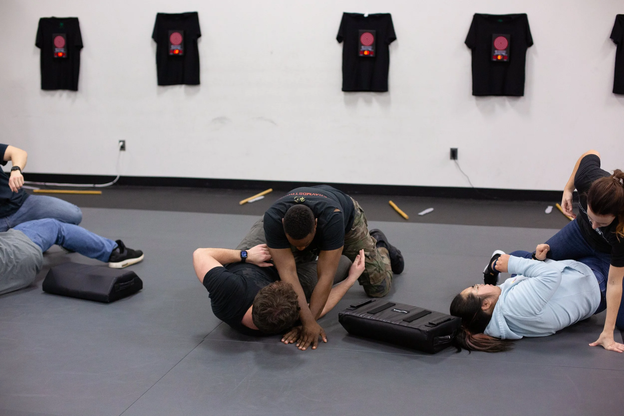 People practicing self-defense techniques on a padded floor in a gym, with wall-mounted black shirts featuring target designs.