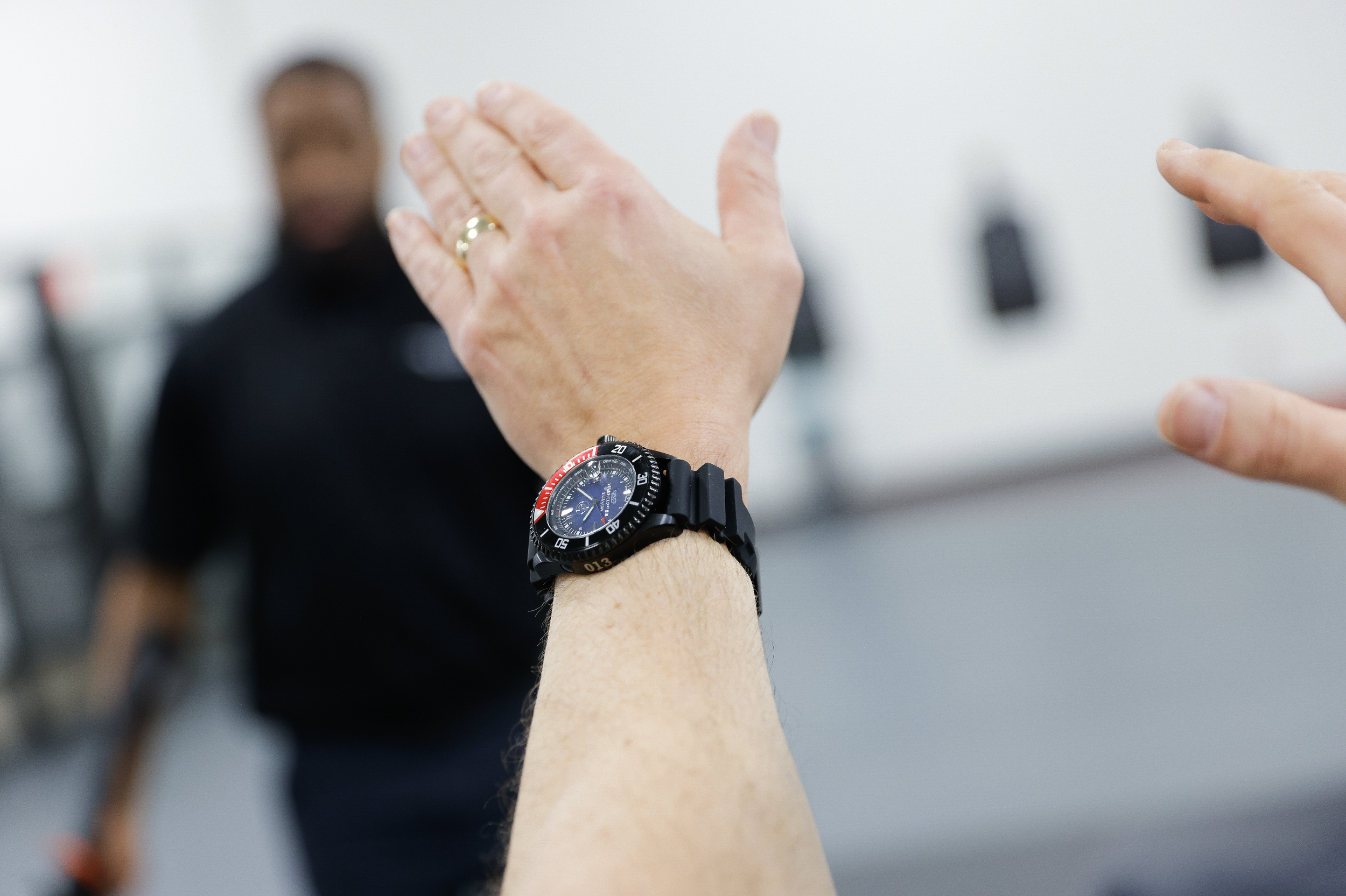Close-up of a person wearing a black wristwatch making a gesture with their hand, with a blurred individual in a black uniform in the background.