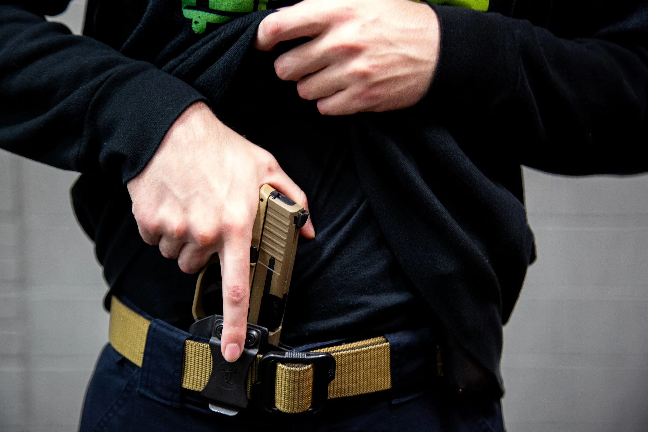 Person drawing a handgun from a holster under a black shirt, wearing a tan belt.