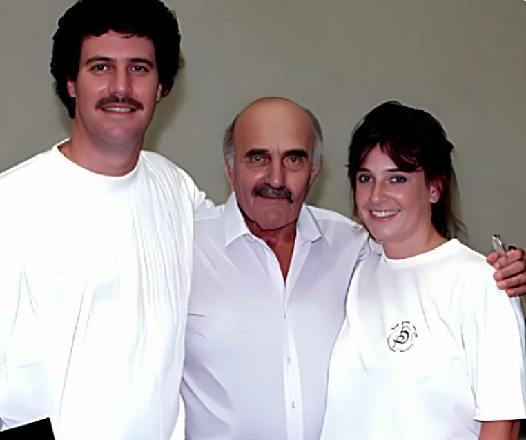 Three people (Darren Levine, Imi Lichtenfeld, Marni Levine) smiling and posing together, two women and one man, wearing white shirts, standing against a beige wall.