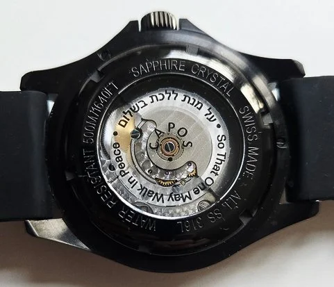 Back view of a black wristwatch showing its mechanical movement, with inscriptions about materials and manufacturing details.