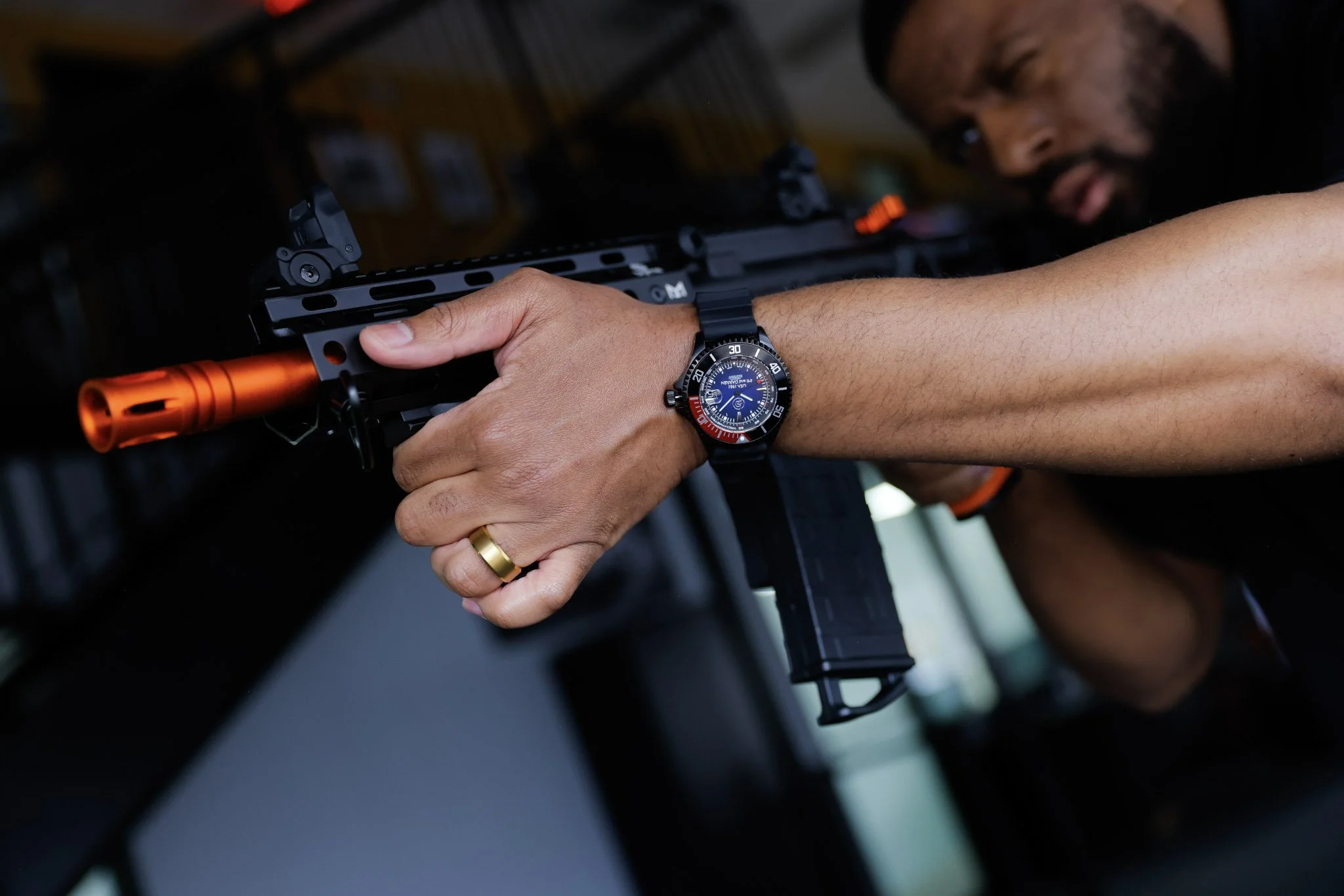 A person aiming a (Byrna) rifle with a red-orange barrel, wearing a wristwatch with a black strap.