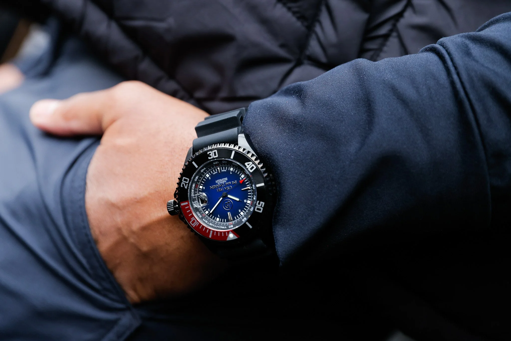 A person wearing a black wristwatch with a blue and red bezel, black strap, and blue dial. The person's hand is resting on their lap, wearing a dark jacket with a zippered sleeve.